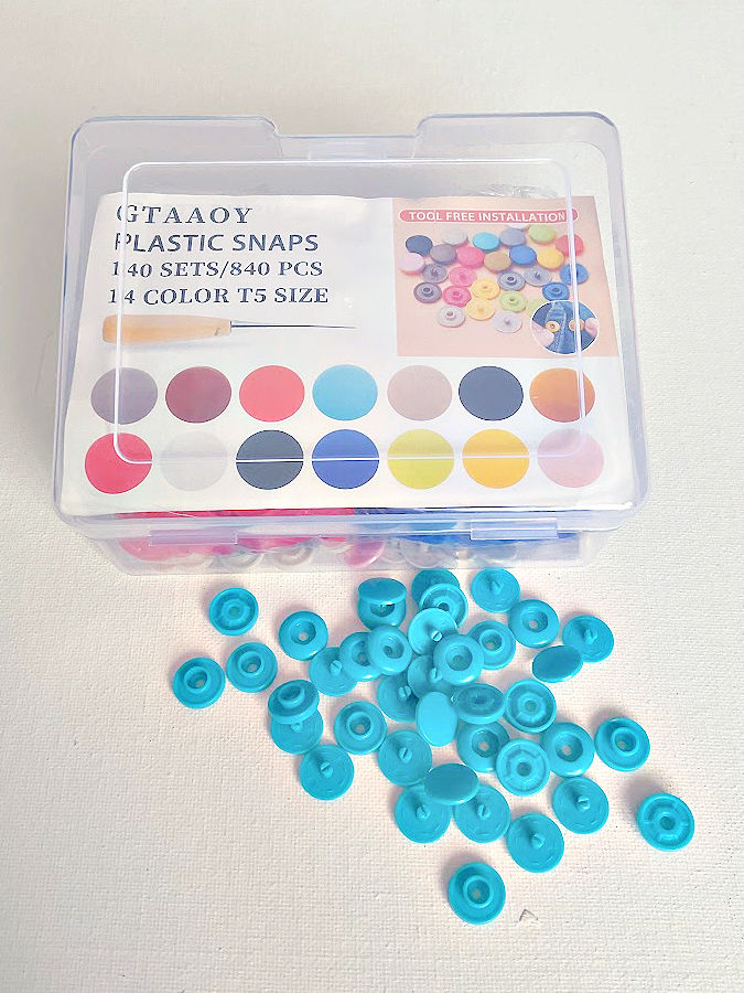 the box of plastic snaps with part of its contents outside the box on the table the label visible on the box reads gtaaoy plastic snaps 140 sets 840 pieces 14 color t5 size