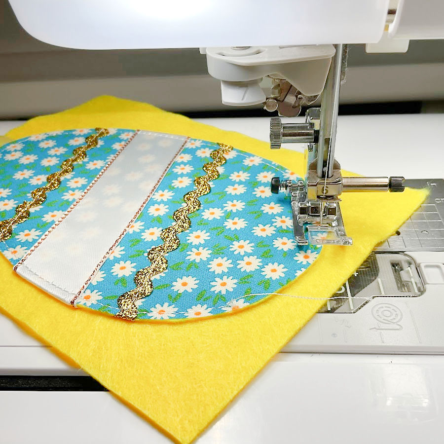 a sewing machine needle assembly shown while sewing the face of the egg to the piece of felt that will be its back the opening for inserting stuffing is visible