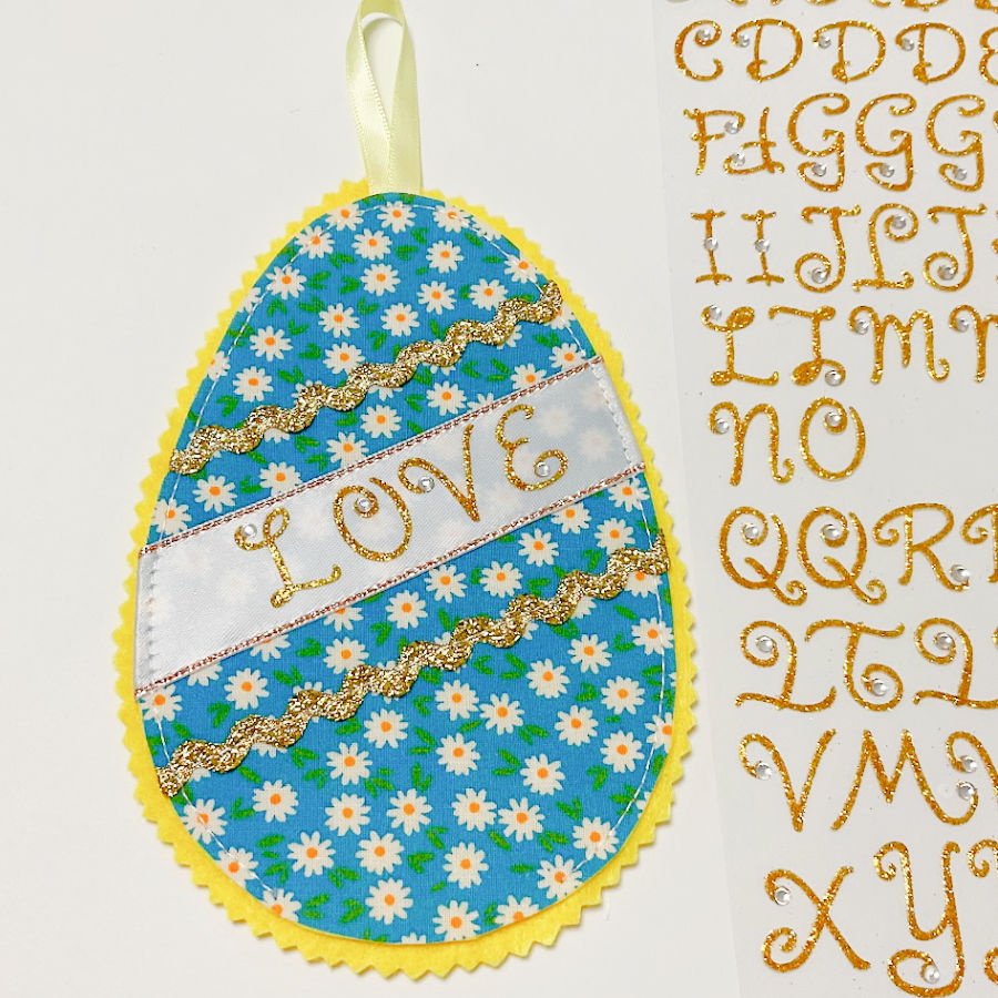 the face of the fabric egg with the word love spelled in golden letters on the wide ribbon a sheet of sticker letters at the right of the image