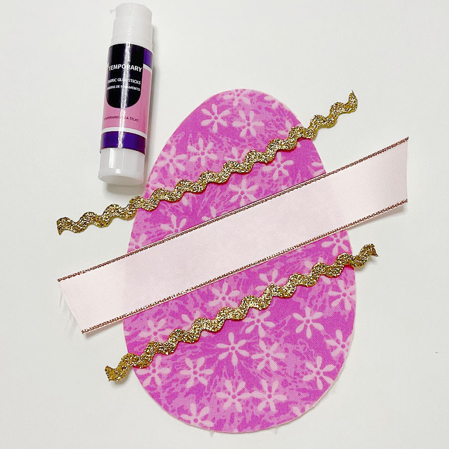 the easter fabric egg face cut on top two pieces of golden decorative ribbon and a piece of white ribbon a stick of fabric glue near