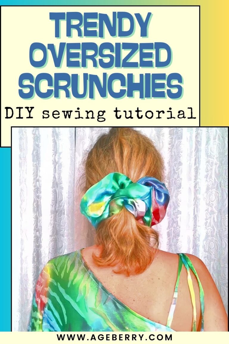 A woman is shown from behind wearing a large, oversized satin scrunchie in a low ponytail. The scrunchie features bright blended colors including green, blue, pink, and yellow with a glossy sheen. She has shoulder-length blonde hair and is wearing a green patterned top with thin straps. The background consists of light-colored curtains, softly lit.