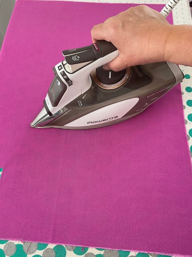a womans hand holding an iron while pressing a piece of colored fabric a wrinkle visible on the fanric