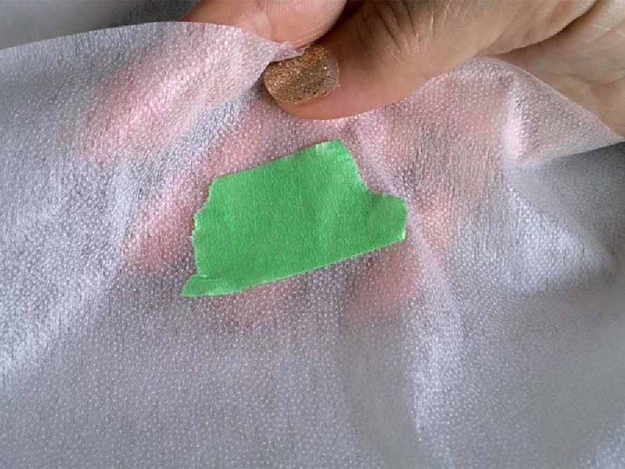 a womans hand showing a piece of interfacing fabric with the glue side marked with piece of green painters tape
