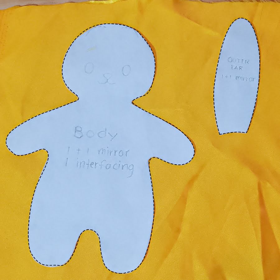 Soft toy body pattern and outer ear pattern placed on yellow fabric, marked with dashed seam lines and notes showing mirrored cuts and interfacing placement.