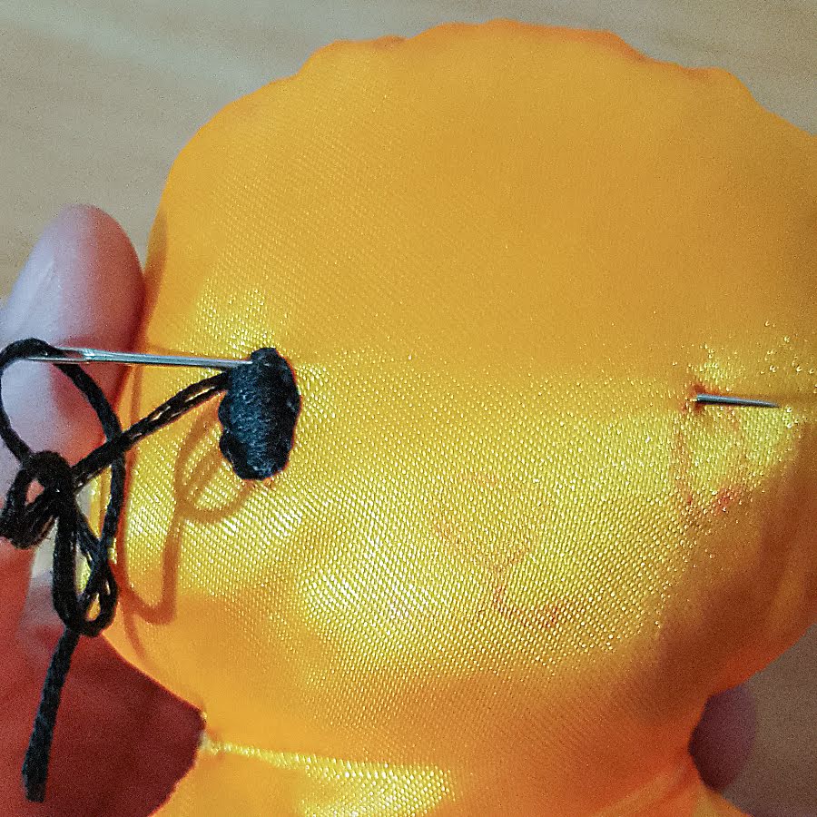 Soft toy face during embroidery of the second eye, showing satin stitch filling being worked with black thread while the needle emerges at the marked eye position to match the first eye.