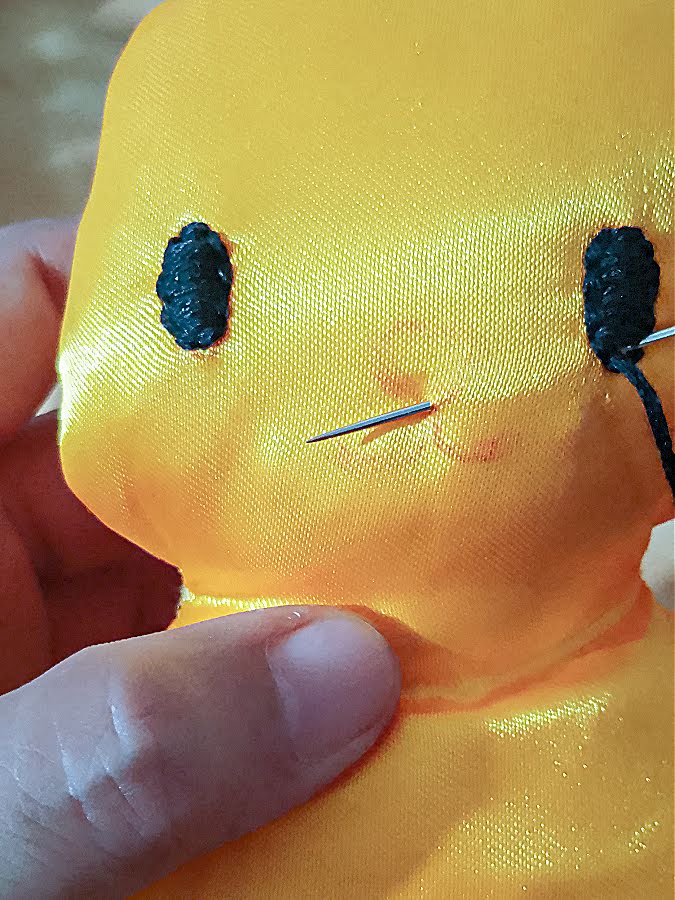 Close-up of mouth embroidery in progress on the soft toy face, with a needle and black thread following the marked mouth line using small, evenly spaced backstitches.