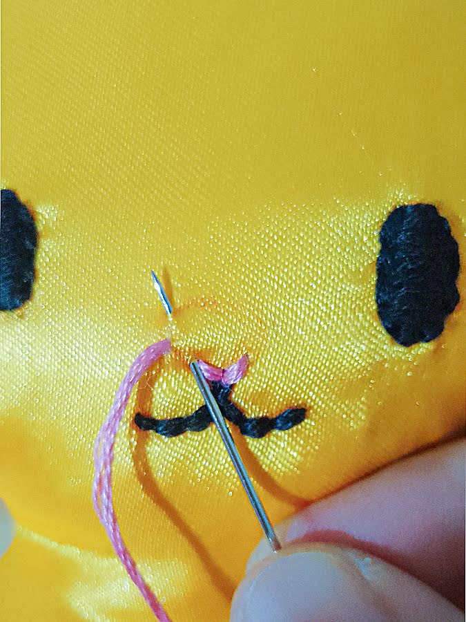 Close-up of the soft toy face as the needle and pink embroidery thread are placed at the marked nose area, beginning the first stitch to anchor the thread before filling the nose.