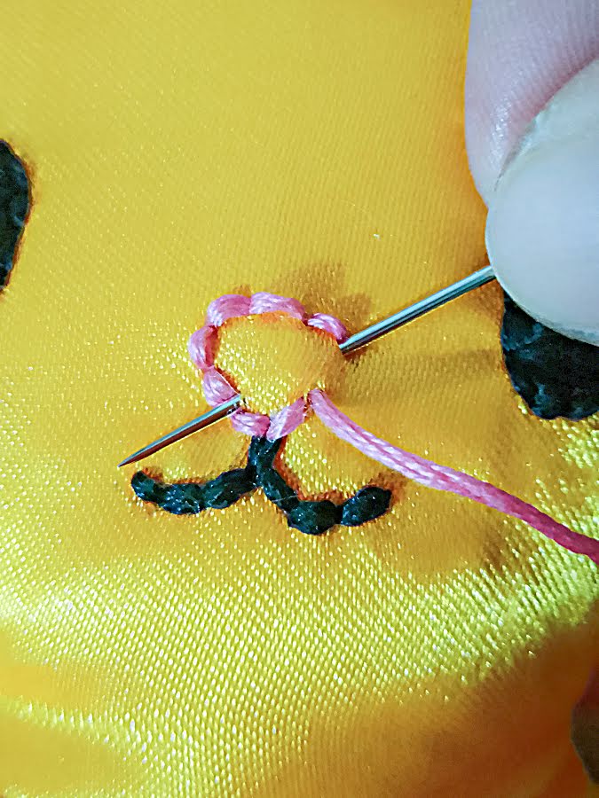 Close-up of the soft toy face showing the nose outline partially stitched with pink thread, as the needle passes through to continue building the edge stitches before filling the nose.