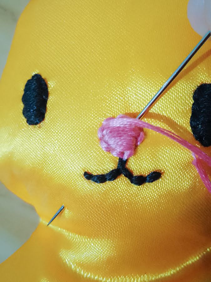 Soft toy face with the nose nearly filled using satin stitch, showing pink embroidery thread laid densely to cover the marked nose shape while the needle completes the final stitches.