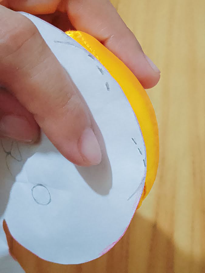 Soft toy head with paper pattern held in place, showing ear placement marks being transferred from the pattern to the fabric to ensure even, symmetrical positioning before attaching the ears.