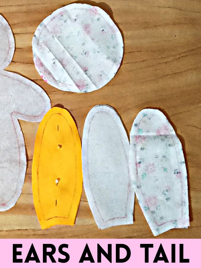 Cut fabric pieces for a soft toy, including outer ear, inner ear, fusible interfacing ear layers, and a circular tail piece, laid out on a wooden surface and ready for assembly.