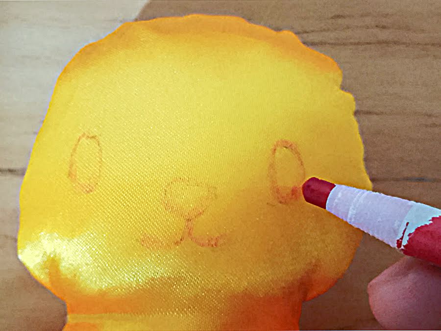 Soft toy head with facial features being drawn on using a fabric marker, showing placement of eyes and nose before stitching or embroidery.