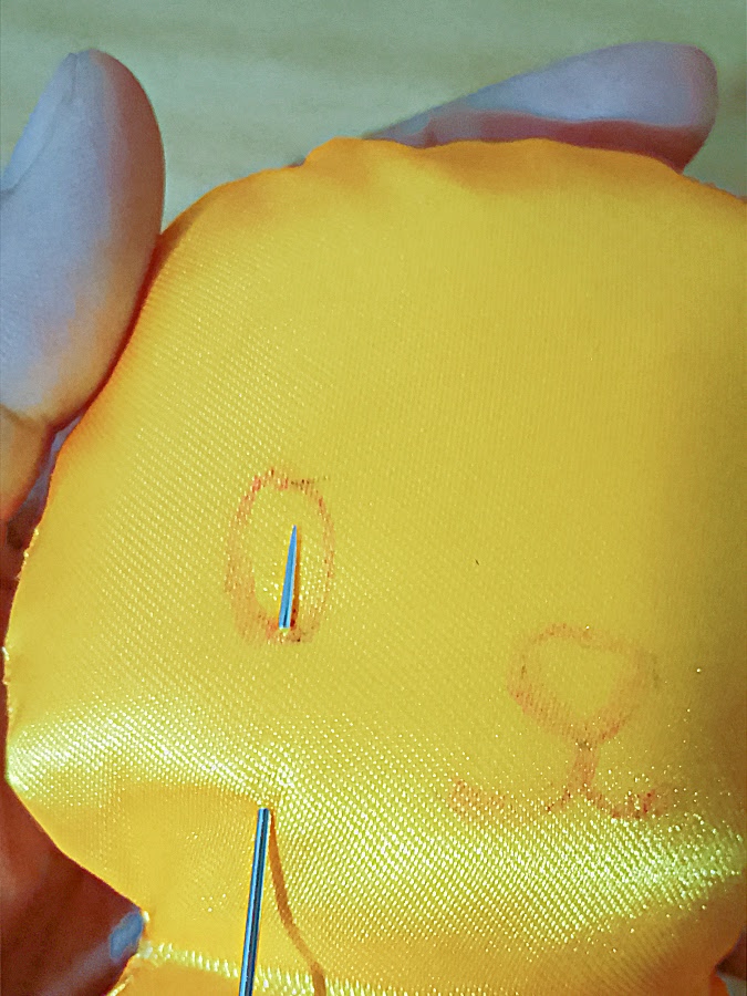 Close-up of the soft toy head with a needle inserted along the drawn eye outline, beginning hand stitching to secure the facial features.