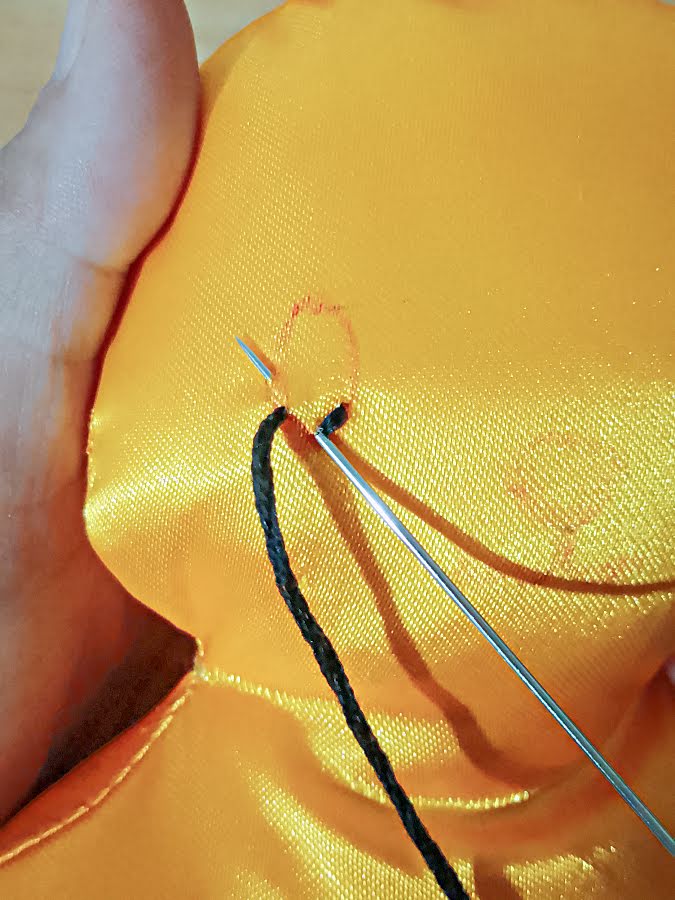 Close-up of hand embroidery on the soft toy face, showing black thread being stitched along the drawn eye shape as the needle passes through the fabric.