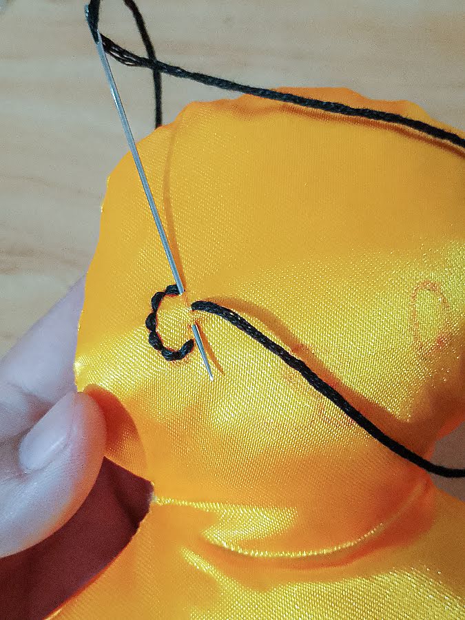 Close-up of soft toy facial embroidery in progress, showing black thread stitched into a small curved eye shape as the needle is pulled through to form tight, even stitches.