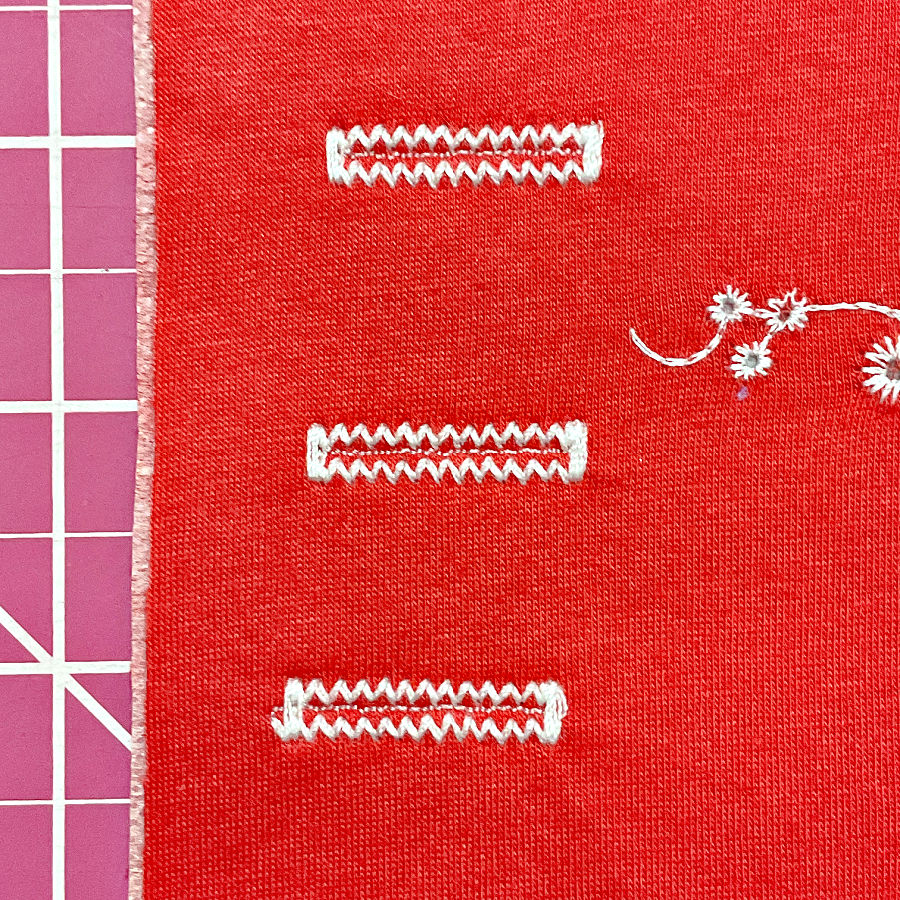 Red knit fabric sample showing three white stretch buttonholes sewn with zigzag stitches.