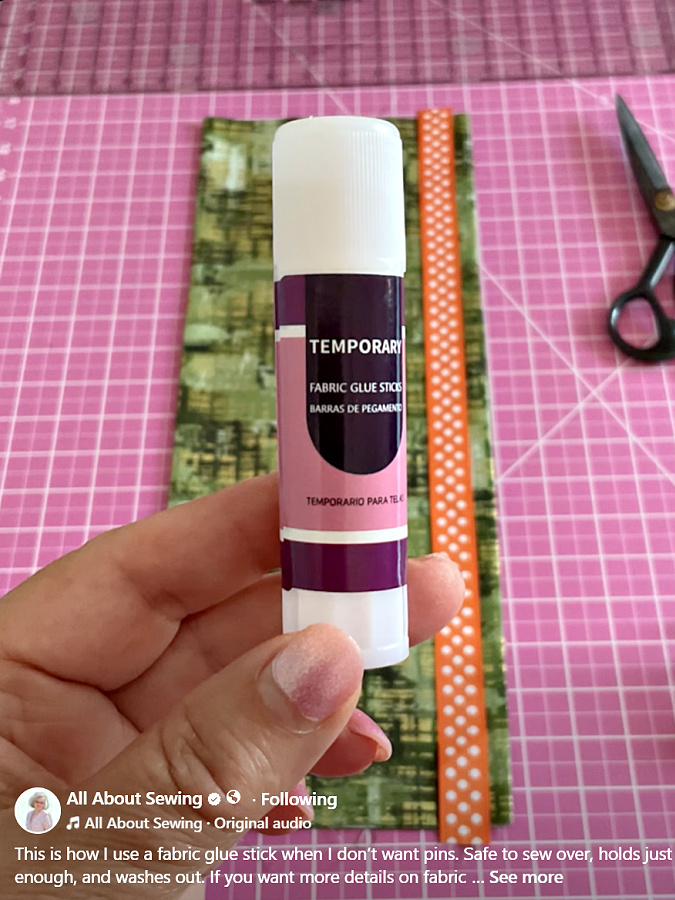 a womans hand holding a fabric glue stick in the background a sewing table with material and scissors on it