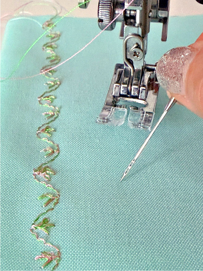 a womans hand holding a double eye needle a sewing machine foot assembly and a decorative stitch in the background