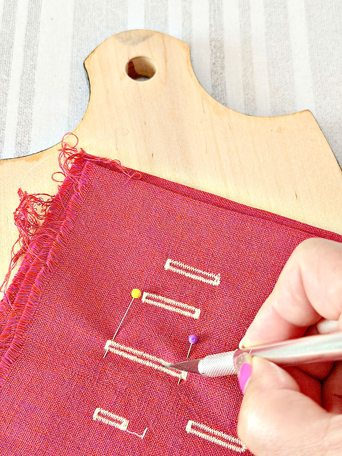 a wooden board sits under a piece of fabric with five buttonholes a womans hand with a knive while cutting the fabric