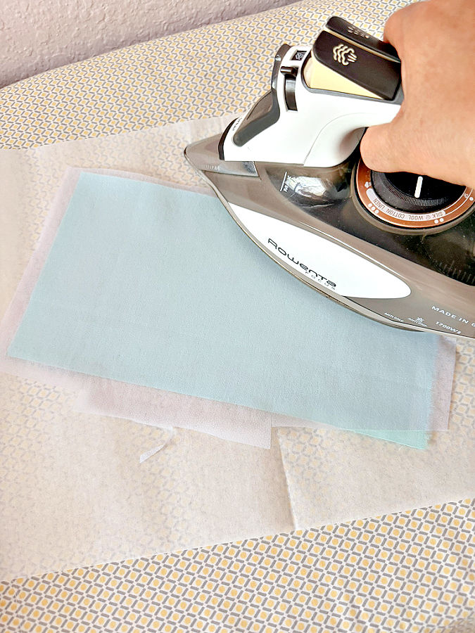 a womans hand holds a hot iron while ironing a piece of interfacing on a piece of fabric under the material a sheet of parchment paper