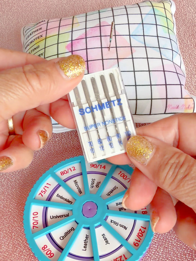 a wom,ans hand holding a pack of schmetz non stiock needles the needle noter and needle storage pillow in the background