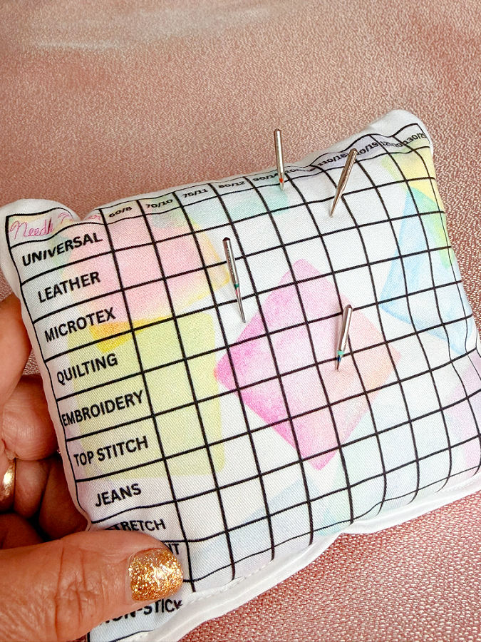a womans hand holds the needle storage pillow for display some needles inserted in the pillow in the locations corresponding to their type