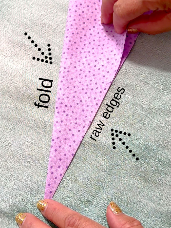 a womans hands shown while folding the fabric in half arrows point to the fold line and the raw edges that need to be aligned
