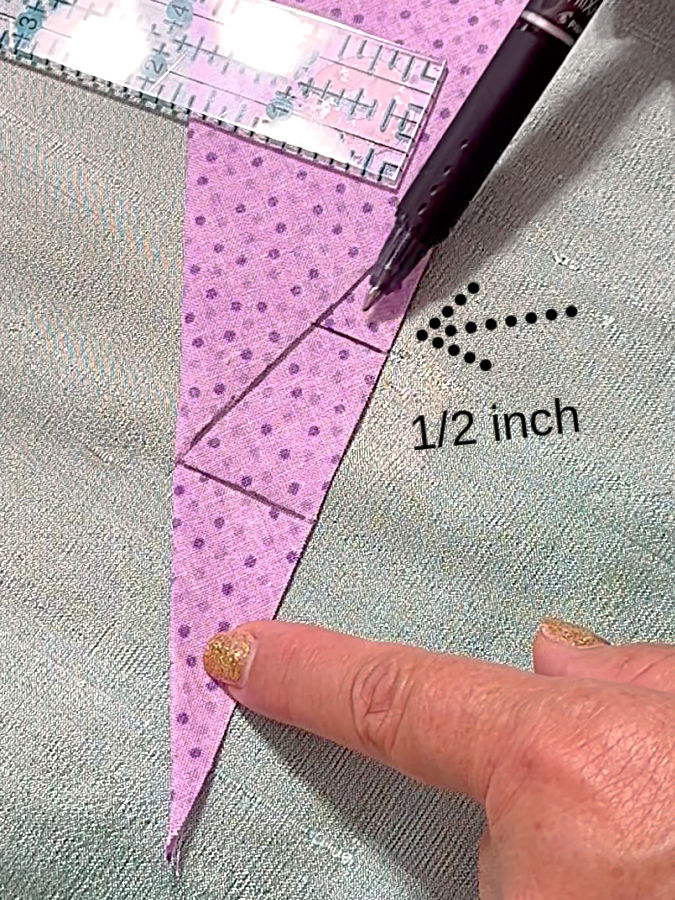 a woman holds down the corner of the fabric an arrow points to the 1/2 inch long line marked before a ruler and pen also in the image