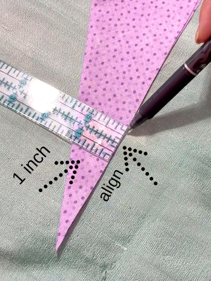 a womans hand holding a pen while drawing the first reference line on the fabric according to the computations described a ruler in the image two arrows point to the 1 inch line and to the edges that need to be aligned