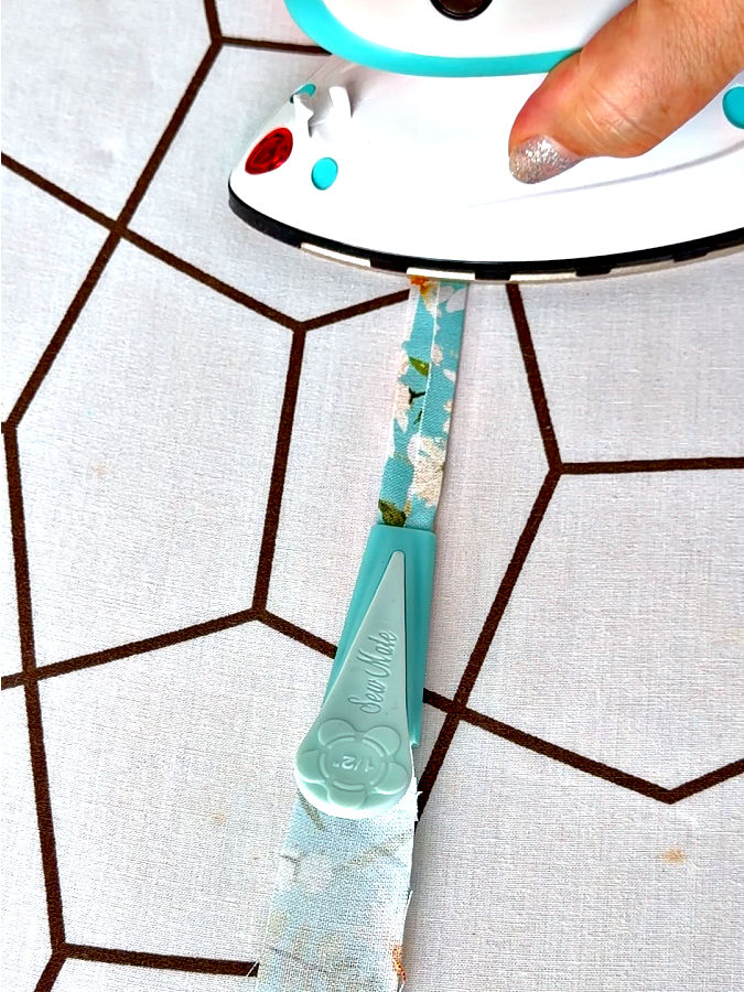a woman uses a mini iron to press the bias tape as it exits the bias tape maker