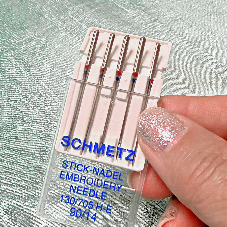 a womans hand holding a pack of schmetz embroidery needles