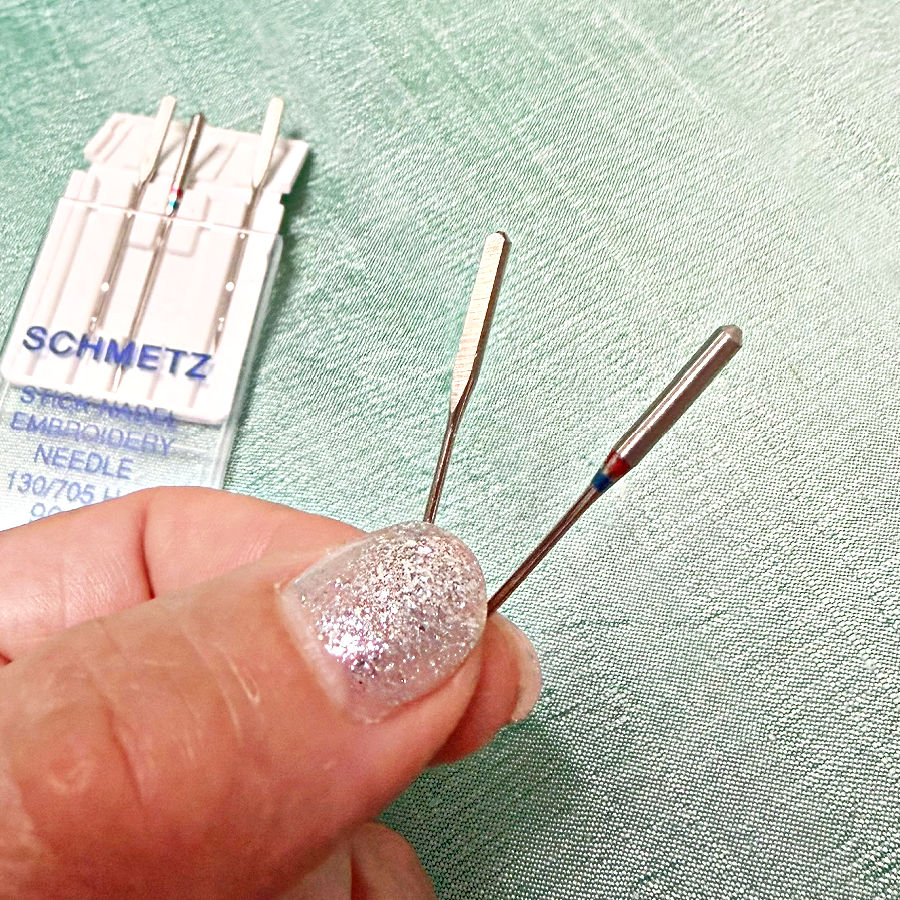 a womans hand holding two sewing machine needles the flat upper part visible on one of the needles a pack of needles in the background
