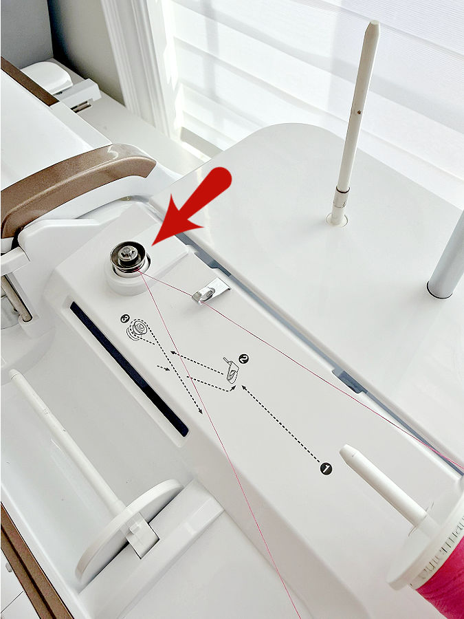the top of a sewing machine showing the threading diagram for the bobbin winder an arrow points to the tension disk