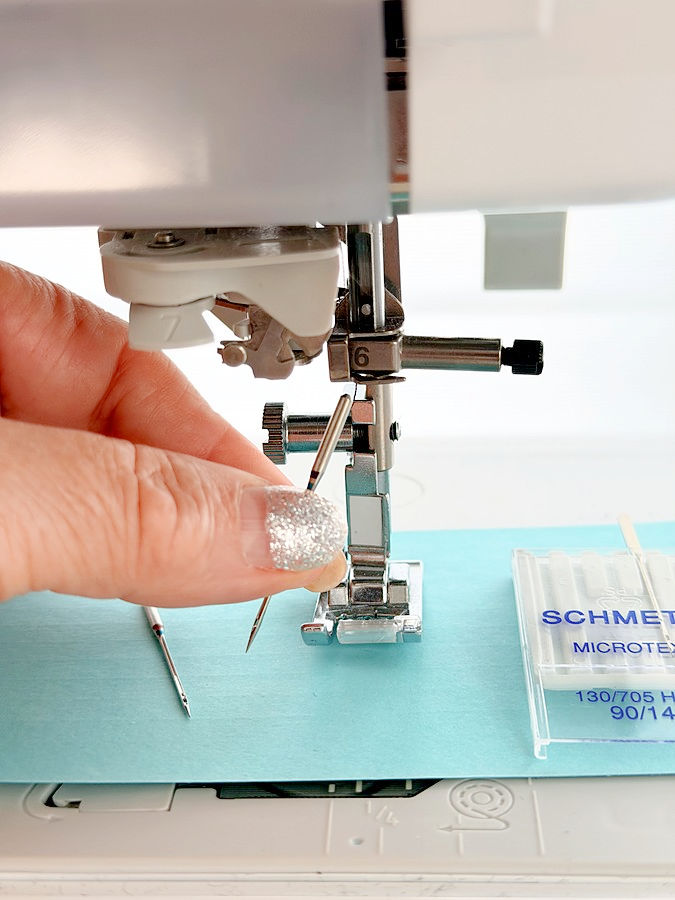 a woman holds between her fingers a needle for a sewing machine the presser foot assembly of the machine in the background